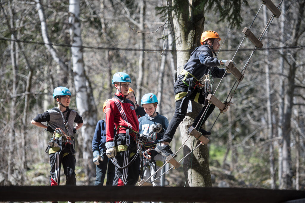 Sechs Kinder mit Helmen in einem Kletterpark, ein Klnd klettert eine Hängeleiter hoch.