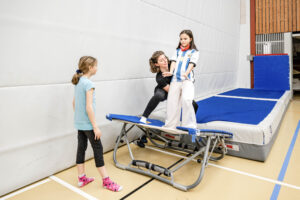 Leiterin hilft einem Kind auf dem Trampolin im KIDS Kurs, ein Mädchen schaut zu