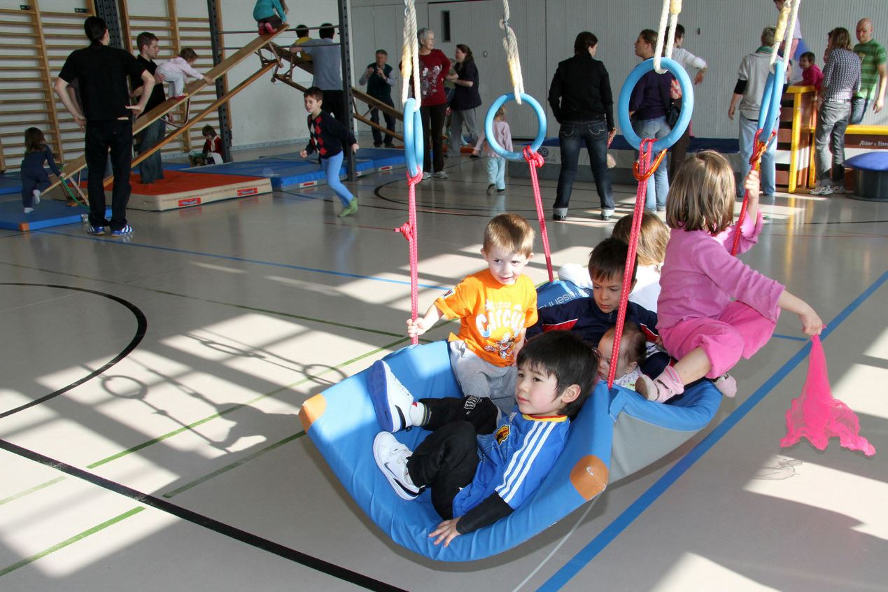 Kinder spielen in Turnhalle auf einer Matte, die an Ringen aufgehängt ist