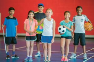 Eine Gruppe Kinder mit Sportgeräten wie Bällen und Schläger in einer Turnhalle